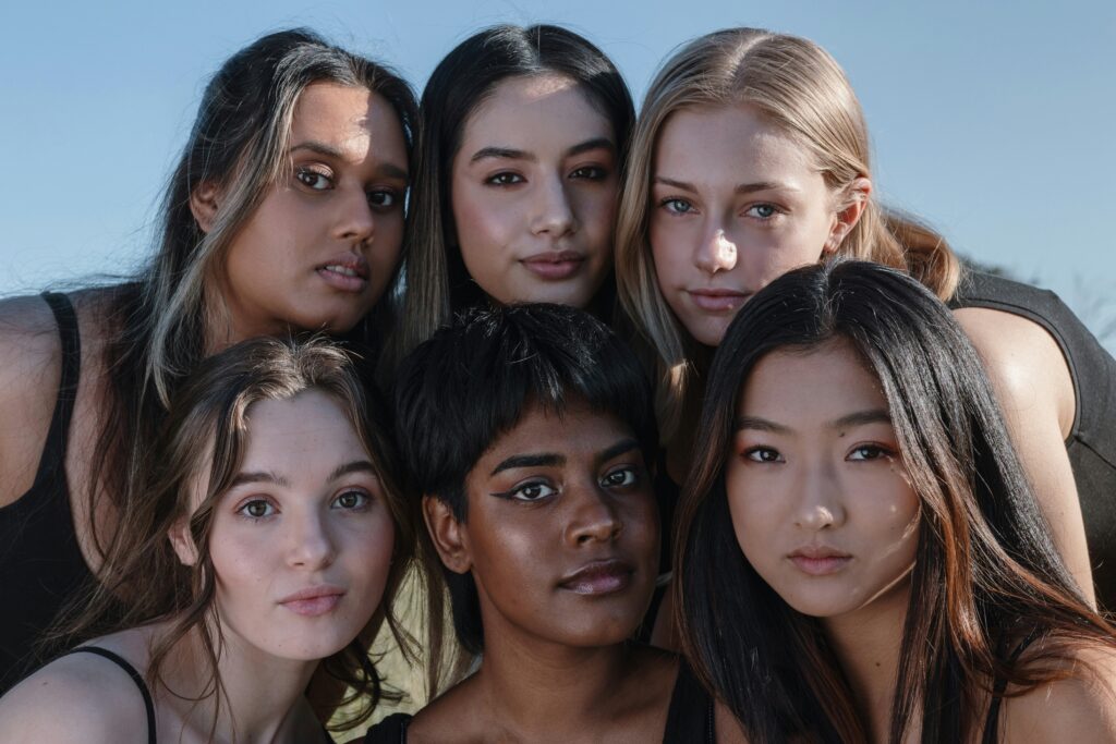 "Diverse group of young women of different ethnicities posing closely together outdoors under a blue sky, smiling confidently at the camera, highlighting friendship, diversity, inclusion, and female empowerment"