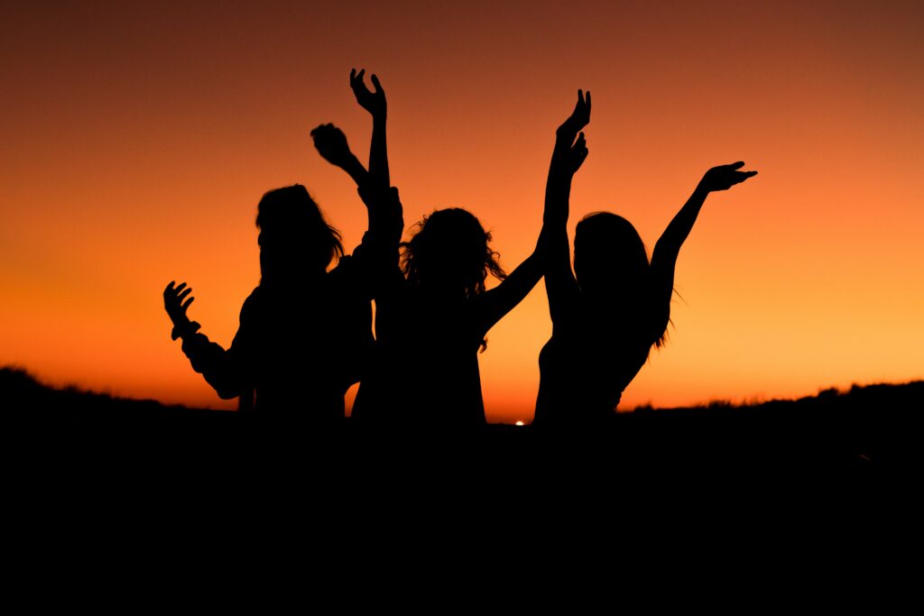 "Silhouette of a group of joyful women jumping and raising their arms in celebration against a vibrant orange sunset sky, symbolizing freedom, empowerment, unity, and female strength"