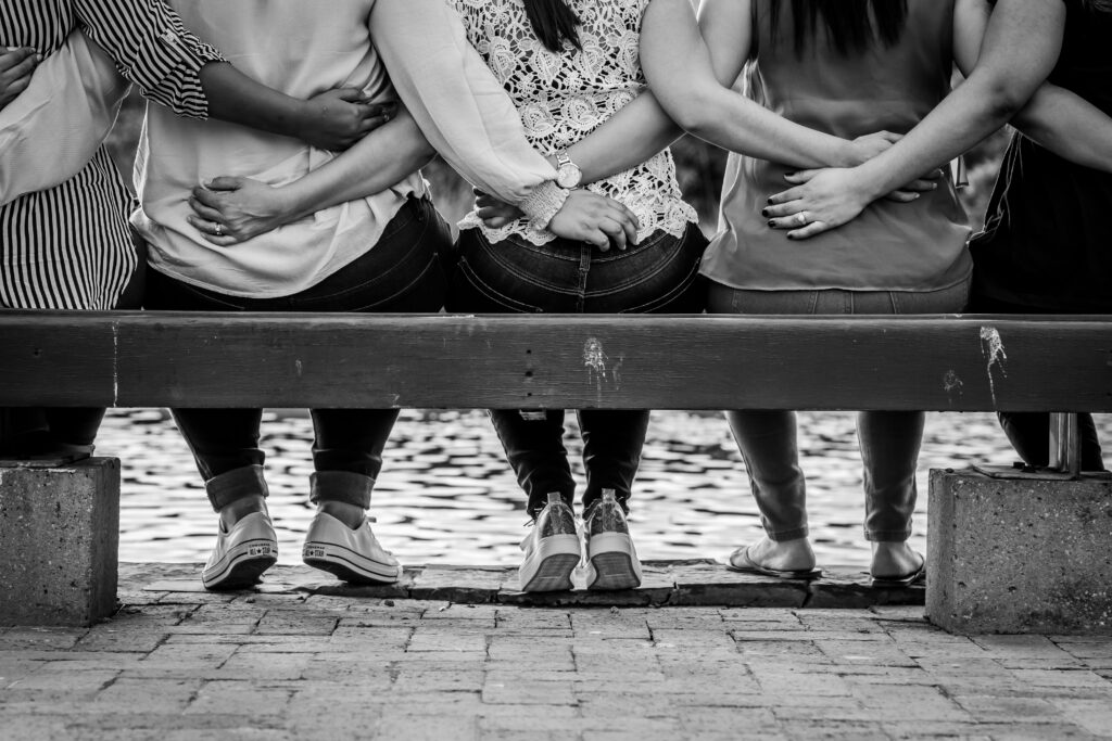 "Black and white photo of three women sitting on a wooden bench with arms linked around each other, symbolizing female solidarity, unity, and empowerment"